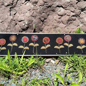 May include: A black felt wall hanging featuring a row of 10 colourful felt flowers. Each flower has a yellow stem and leaves. The flowers are arranged in a row against a black background.