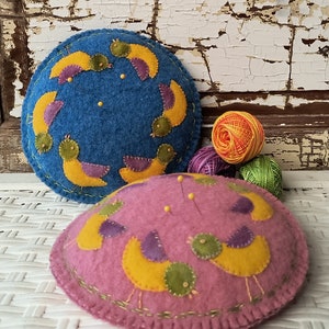 May include: Two felt pincushions in the shape of a circle with birds embroidered on them. The pincushion on the left is blue with yellow birds and the pincushion on the right is pink with yellow birds. Both pincushions have green accents.