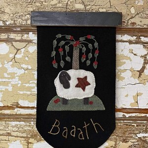 Puede incluir: Una pancarta de fieltro negro con una oveja blanca de pie sobre una colina verde. La oveja tiene una estrella roja en un lado y un sauce llorón con bayas rojas encima. La pancarta tiene la palabra "Baath" bordada en hilo dorado.