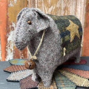 Puede incluir: Figura de oveja gris artesanal con una campana alrededor del cuello y un diseño de estrella en la espalda. La oveja está hecha de lana afieltrada y se apoya sobre una base decorativa con detalles de tela colorida. Decoración rústica, estilo campestre.