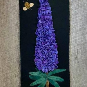 May include: A purple flower with green leaves and a bee on a black background. The flower is made of felt and is a wall hanging.
