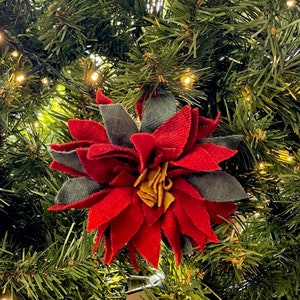 Peut inclure: Une décoration de Noël en forme d'étoile de Noël en feutre rouge avec des feuilles en feutre vert, suspendue à un sapin de Noël artificiel vert.