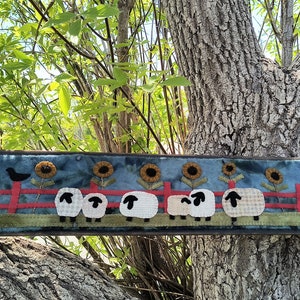 May include: A blue and green fabric wall hanging with a red fence, white sheep, and black birds. The sheep are lined up in a row and there are sunflowers behind them.