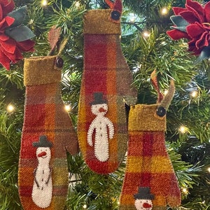 Peut inclure: Trois décorations de mitaines en feutre avec des motifs de bonhomme de neige accrochés à un sapin de Noël. Les mitaines sont en tissu écossais rouge, vert et jaune avec des bonhommes de neige en feutre blanc.