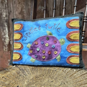 以下が含まれることがあります： 手縫いの斑点のある亀のデザインが施された青と黄色の枕。枕には赤い刺繍で「Spotted Turtle」と書かれています。
