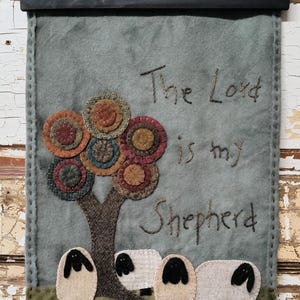 May include: A blue felt wall hanging with a black wooden frame. The hanging features a colorful tree with the text "The Lord is my Shepherd" embroidered on it. Three white sheep with black faces are standing below the tree.