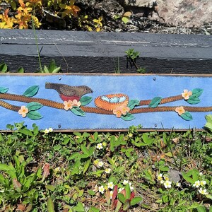 May include: A blue felt wall hanging with a brown bird, a nest with blue eggs, and orange flowers on a brown branch.