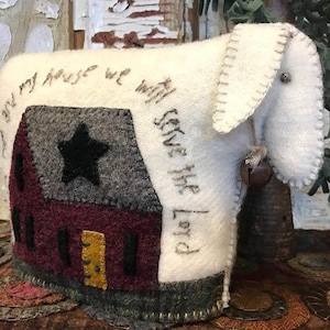Puede incluir: Un cordero de fieltro blanco con una casa roja, gris y verde en su lado. La casa tiene una estrella negra en el techo y el texto "And in my house we will serve the Lord" está bordado en el lado del cordero.