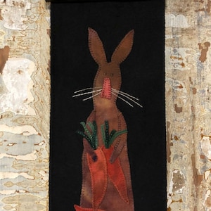 May include: A brown felt bunny with a pink nose holding a bunch of orange carrots on a black felt background. The bunny is surrounded by green felt leaves. The felt piece is framed with a black wooden frame.