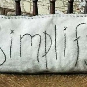 Puede incluir: Una almohada blanca con costuras negras y la palabra "Simplificar" bordada en hilo negro.