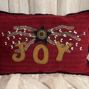 May include: A red wool pillow with a black border and a white and green star with a button in the center. The word "JOY" is embroidered in gold yarn with white sprigs around the star.