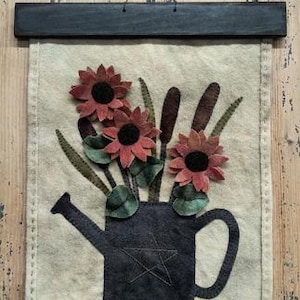 Peut inclure: Une tenture murale avec un motif en feutre de fleurs dans un arrosoir. Les fleurs sont rouges avec des centres noirs, et l'arrosoir est bleu foncé. Le fond est de couleur beige clair, et la pièce est encadrée d'une barre en bois foncé.