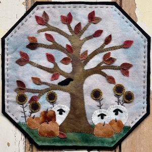 Peut inclure: Applique en feutre octogonal représentant un arbre avec des feuilles rouges et marron, un oiseau noir et des tournesols. L'arbre est centré sur un fond bleu clair, avec des citrouilles marron et des moutons blancs à la base. Le tout est encadré d'une bordure noire.