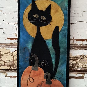 May include: A black felt cat sitting on two orange felt pumpkins in front of a yellow felt moon. The background is a blue felt with a green and blue ombre effect.