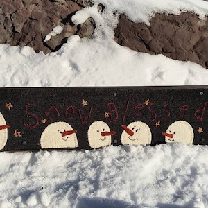Könnte beinhalten: Ein schwarzes Filzschild mit den Worten "Snow Blessed" in roter Stickerei. Fünf weiße Filz-Schneemänner mit orangenen Nasen und schwarzen Augen sind über das Schild verteilt.