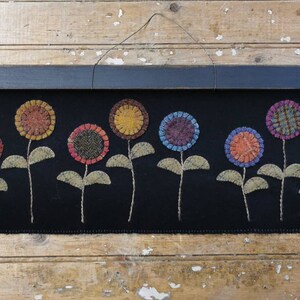 May include: A black fabric wall hanging with a row of colorful, embroidered flowers. Each flower has a patterned center and is attached to a stem with green leaves. The hanging is topped with a dark wooden bar.
