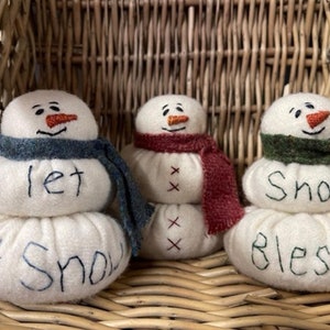 May include: Three white felt snowman ornaments with embroidered faces and scarves. The snowmen are stacked in three tiers and have the words "Let it Snow", "Snow" and "Snow Blessed" embroidered on them.