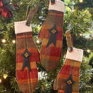 May include: Three red, green, and brown plaid mitten ornaments with a brown button and a black felt star on each. The ornaments are hanging on a green Christmas tree.