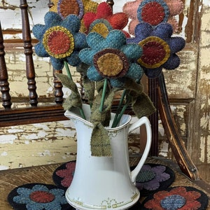 Peut inclure: Un bouquet de fleurs en feutre dans des tons de bleu, rose, rouge et marron, disposé dans un pichet blanc. Les fleurs sont posées sur une surface en bois avec cinq dessous-de-verre en feutre en forme de fleurs.