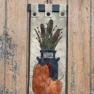 May include: A rectangular wall hanging featuring a milk jug with cattails and pumpkins. The background is a light beige fabric with a dark border. The pumpkins are orange, and the milk jug is dark blue. The hanging has a dark blue top with buttons.