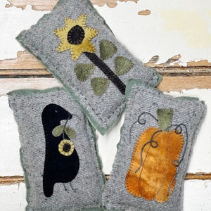May include: Three grey felt cushions with embroidered designs. One cushion features a yellow sunflower, another a black bird holding a sunflower, and the third a brown pumpkin.