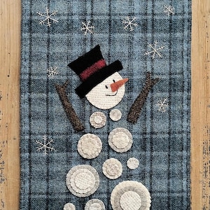 May include: A blue and black plaid fabric wall hanging with a snowman made of white felt circles. The snowman has a black hat with a red band, a white face, and a carrot nose. The background is decorated with white felt snowflakes.