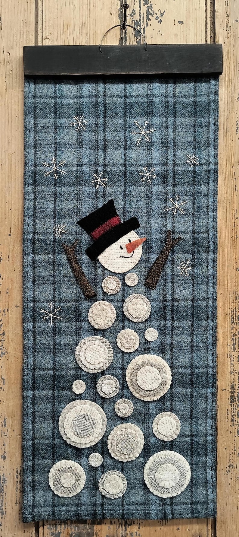 K&ouml;nnte beinhalten: Ein Wandbehang aus blauem und schwarzem kariertem Stoff mit einem Schneemann aus wei&szlig;en Filzkreisen. Der Schneemann tr&auml;gt einen schwarzen Hut mit rotem Band, ein wei&szlig;es Gesicht und eine Karottennase. Der Hintergrund ist mit wei&szlig;en Filzschneeflocken verziert.