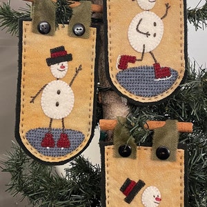 May include: Three felt ornaments featuring a snowman wearing a black hat and red boots. Each ornament is attached to a cinnamon stick and a green felt loop. The ornaments are hanging on a green artificial Christmas tree.