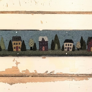 May include: A blue and brown felt wall hanging with a row of colorful houses and trees. The houses are different shapes and sizes and have white windows and doors. The trees are green and have brown trunks.