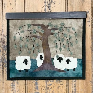 Può includere: Un arazzo da parete in feltro incorniciato con un albero marrone con foglie verdi e tre pecore bianche con facce nere. Lo sfondo è di colore marrone chiaro.