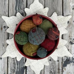 May include: A collection of eight handmade felt baubles in various shades of green, brown, and red, with button accents, displayed in a red bowl on a white snowflake-shaped fabric.