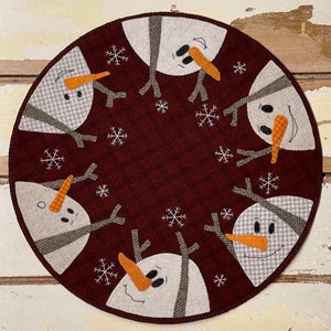 Peut inclure: Un chemin de table rond en feutre bordeaux avec cinq applications de bonhomme de neige en feutre blanc. Chaque bonhomme de neige a des yeux en boutons noirs, un nez de carotte orange et une écharpe en feutre gris. Les bonhommes de neige sont disposés en cercle autour du centre du chemin de table. Le chemin de table est décoré de flocons de neige en feutre blanc.