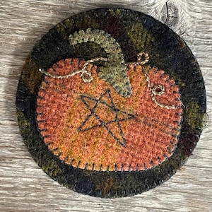 May include: A round wool felt ornament with a stitched design of a pumpkin with a star inside. The pumpkin is orange and brown with a green stem. The background is black with green and brown accents.