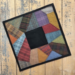 Può includere: Un quilt quadrato a patchwork con un centro nero e un bordo di tessuti di vari colori. Il quilt è fatto di lana e ha un aspetto rustico e fatto a mano.