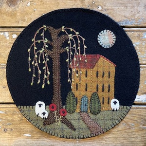 Peut inclure: Une tapisserie ronde en feutre de laine sur fond noir. Une maison jaune avec un toit rouge est au centre de l'image. Un saule pleureur se trouve à gauche de la maison. Deux moutons blancs sont au premier plan. Une pleine lune se trouve dans le coin supérieur droit de l'image.