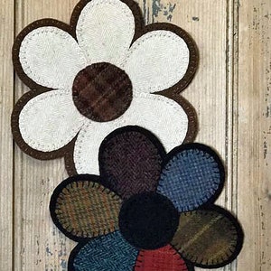 May include: Two felt flower appliques. The top flower is white with a brown center. The bottom flower has petals in shades of brown, blue, and red with a black center.