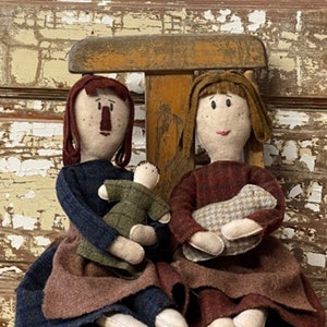 Puede incluir: Dos muñecas de tela hechas a mano sentadas en una silla de madera. La muñeca de la izquierda viste un vestido azul marino y sostiene una muñeca verde más pequeña. La muñeca de la derecha viste un vestido granate y sostiene una manta estampada.
