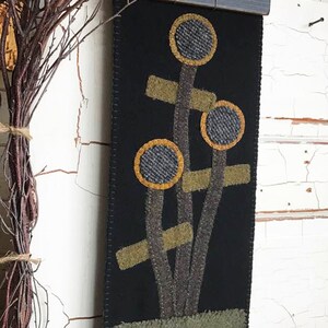 May include: A black felt wall hanging featuring a stylised design of sunflowers in shades of brown, yellow and green. The text "Sunflowers" is embroidered in green at the bottom of the hanging.