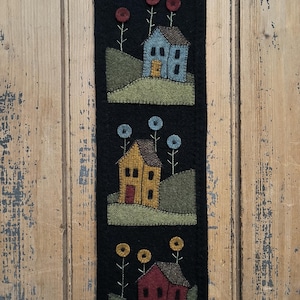 May include: A black felt wall hanging with three rows of houses and flowers. Each row features a different colored house with a green lawn and flowers. The houses are yellow, blue, and red.