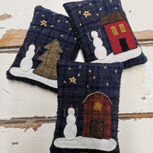 May include: Three blue and white felt ornaments with a snowman, a Christmas tree, and a barn in a snowy night scene. Each ornament has a gold star and a white background.