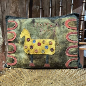 May include: A decorative cushion with a hand-stitched design. The cushion features a yellow pony with button accents and the words "Spotted Pony" embroidered on a mottled green background. Red and green decorative elements frame the design.