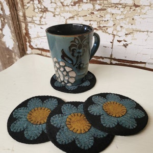 May include: Three blue and yellow felt flower coasters with black backing. The coasters are stacked on a white surface. A blue and black patterned mug is sitting on a coaster.
