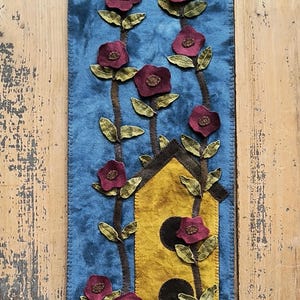 May include: A blue felt wall hanging featuring a yellow birdhouse and red flowers. The birdhouse has three black circles on the front. The flowers are on stems with green leaves.