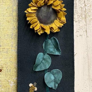 May include: A felt wall hanging with a yellow sunflower and green leaves. A small bee is embroidered below the sunflower.