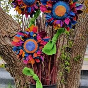 May include: Three colorful fabric sunflowers with vibrant petals in shades of blue, orange, pink, and purple. The flowers have green stems and are arranged in a black metal container. The background features a tree trunk and foliage.