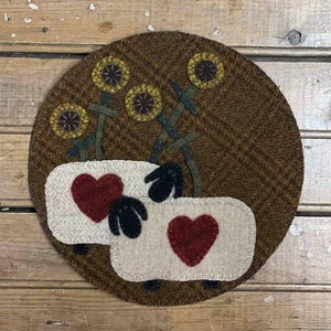 Puede incluir: Un círculo de fieltro de lana marrón con dos ovejas blancas con corazones rojos y caras negras. Las ovejas están rodeadas de girasoles amarillos con tallos verdes.