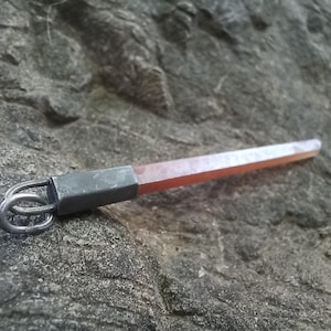 May include: A handcrafted pendant featuring a reddish-orange crystal with a rectangular shape. The crystal is attached to a silver-coloured metal piece with a loop design. The pendant is displayed against a textured grey stone background.