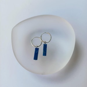 Può includere: Un paio di orecchini a cerchio in argento con pendenti rettangolari in pietra preziosa blu scuro. Gli orecchini sono esposti su un piatto bianco smerigliato di forma organica. Lo sfondo è bianco neutro morbido.