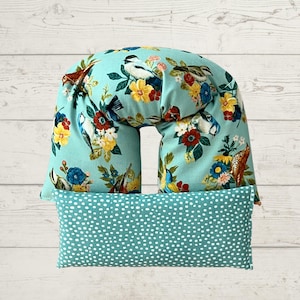 May include: A teal-colored neck pillow and a matching rectangular pillow. The neck pillow features a floral and bird pattern with yellow, red, and blue flowers. The rectangular pillow has a white polka dot pattern.