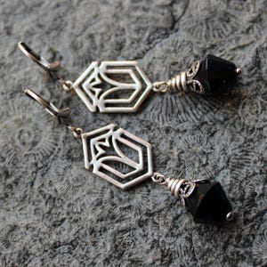 Könnte beinhalten: Ein Paar silberfarbene Ohrhänger. Jeder Ohrring hat ein geometrisches, offenes Design mit floralem Motiv, verbunden mit einem facettierten schwarzen Edelstein. Die Ohrringe haben einen Klappverschluss.